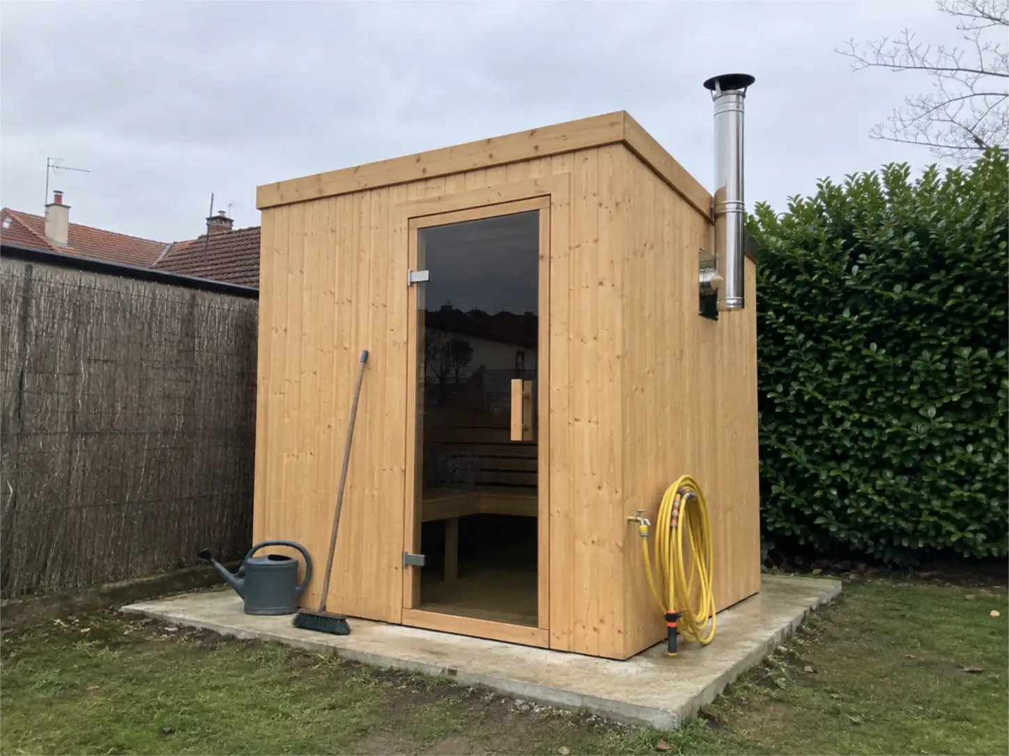 Sauna extérieur en cabine carrée à bardage vertical, installé sur une dalle béton dans un jardin