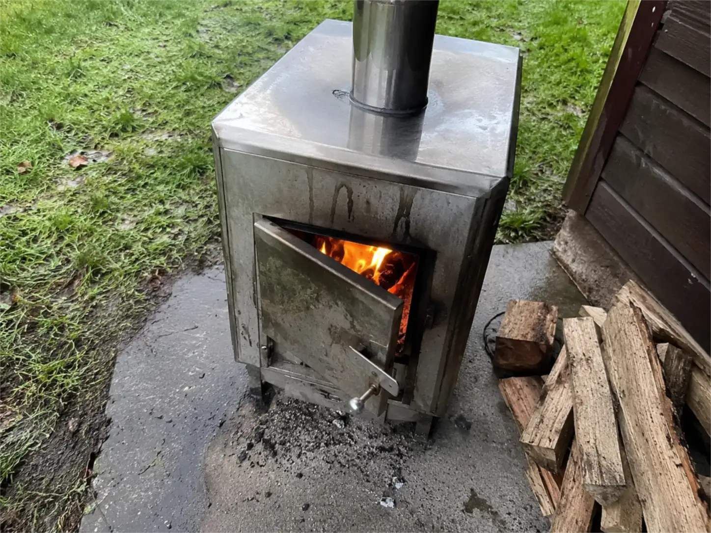 Poêle à bois inox extérieur d'un sauna, porte du foyer entrouverte avec les flammes visibles et bûches à côté