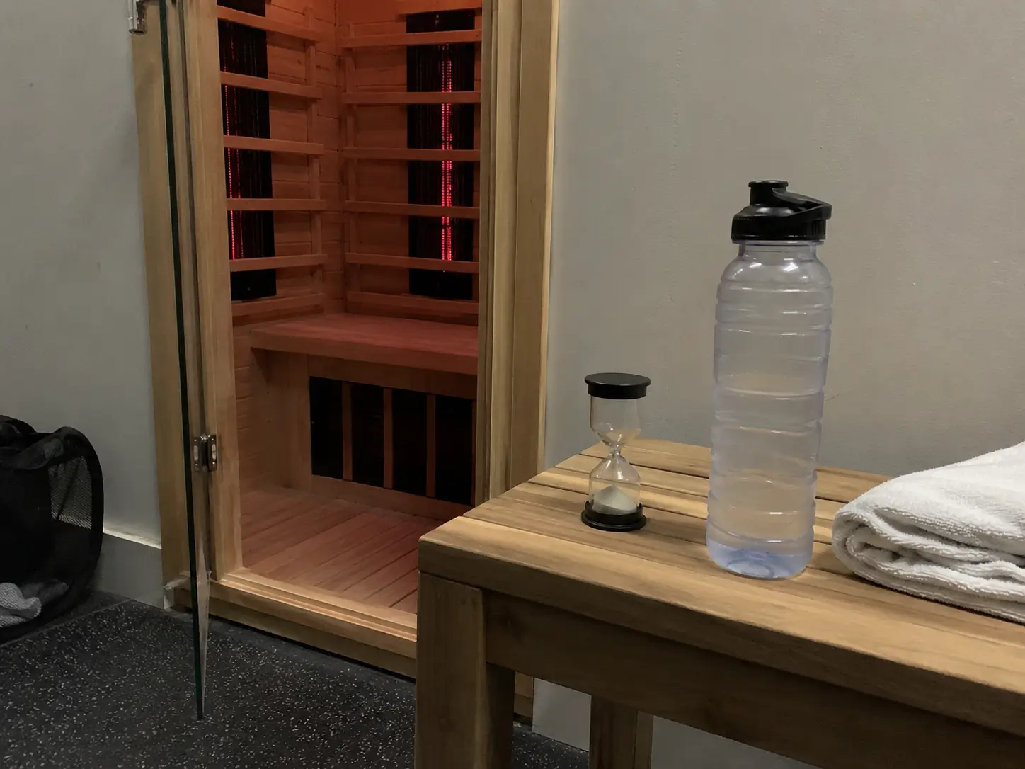 Sablier de 15 minutes et bouteille d’eau posés sur un banc devant une cabine de sauna infrarouge entrouverte