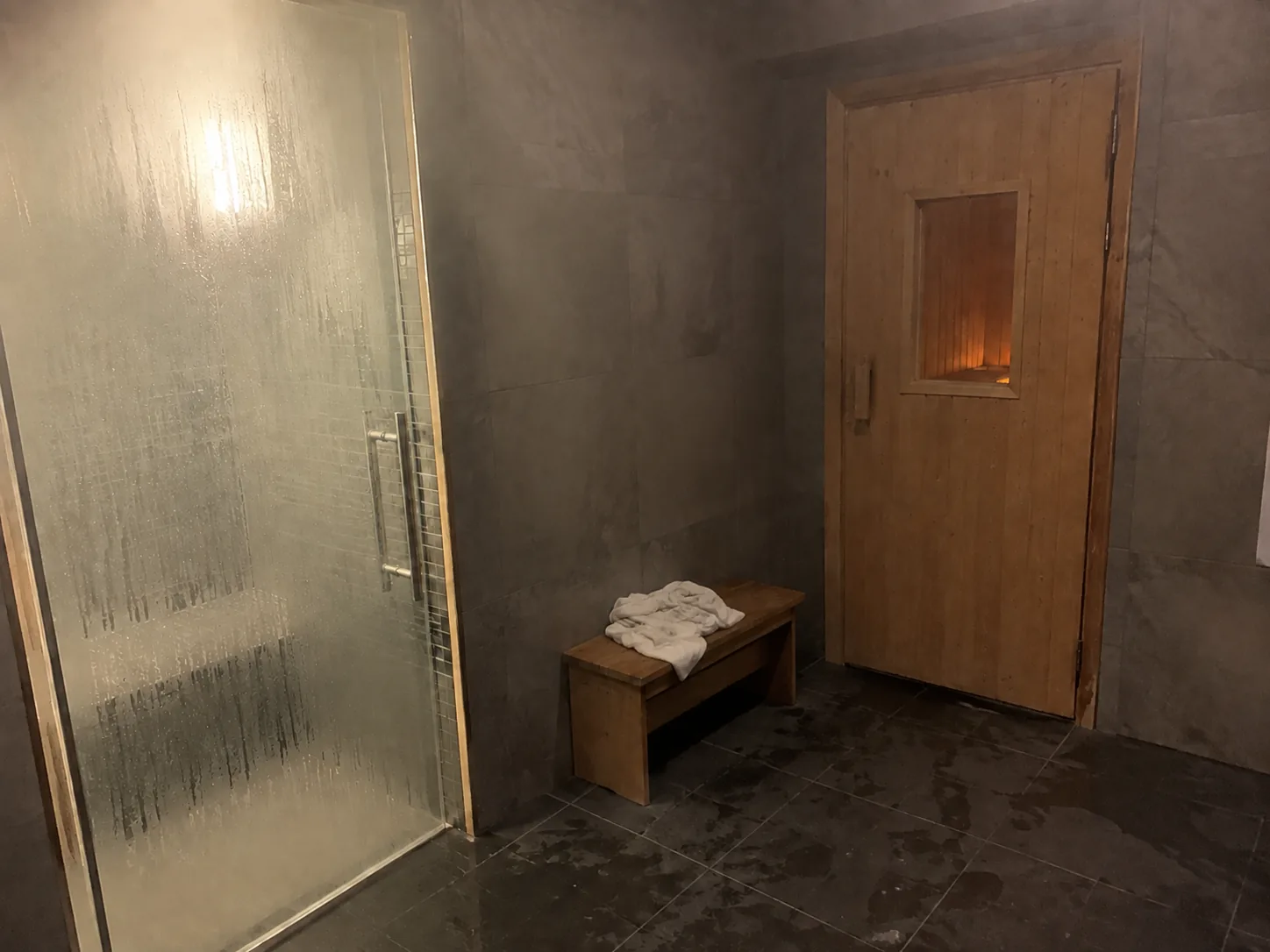 Porte vitrée embuée du hammam à gauche et porte en bois du sauna à droite, serviette blanche sur un banc et carrelage humide