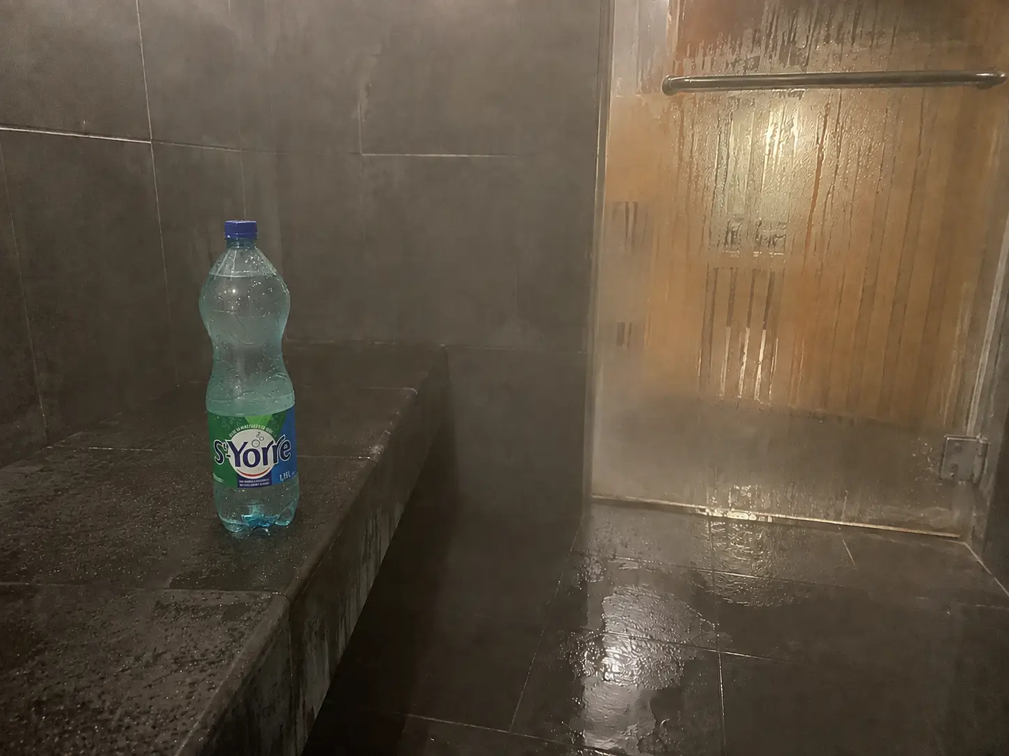 Banc carrelé trempé dans un hammam saturé de vapeur, gouttes d'eau sur la porte vitrée embuée et bois du sauna derrière